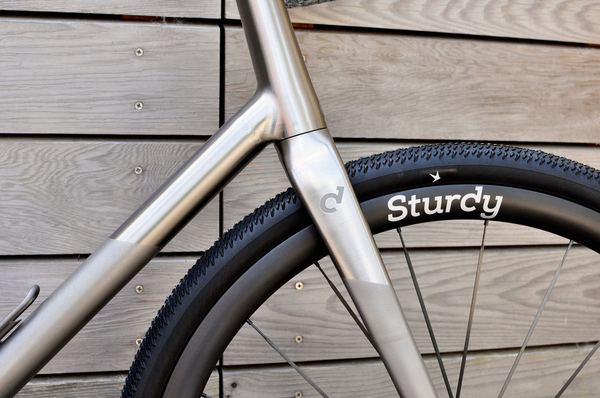 Polished and shot-peened titanium fork and down tube with Sturdy wheel against a grey timber wall