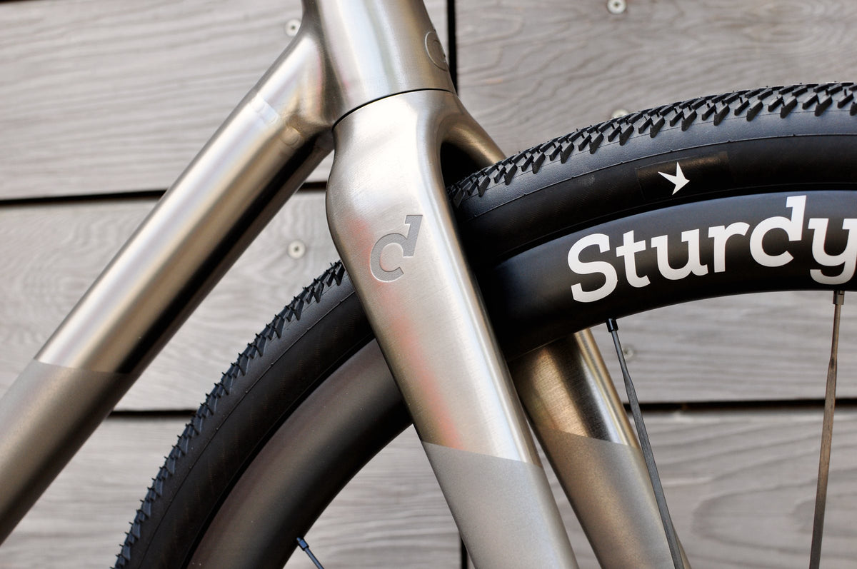 Titanium fork crown with etched logo and polished finish beside a Sturdy wheel and gravel tyre