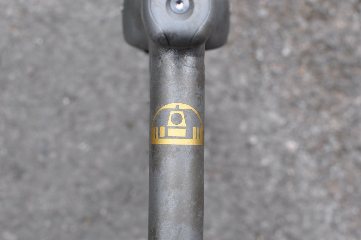 Gold domed robot emblem on a shot-peened titanium top tube, seen from above against grey tarmac