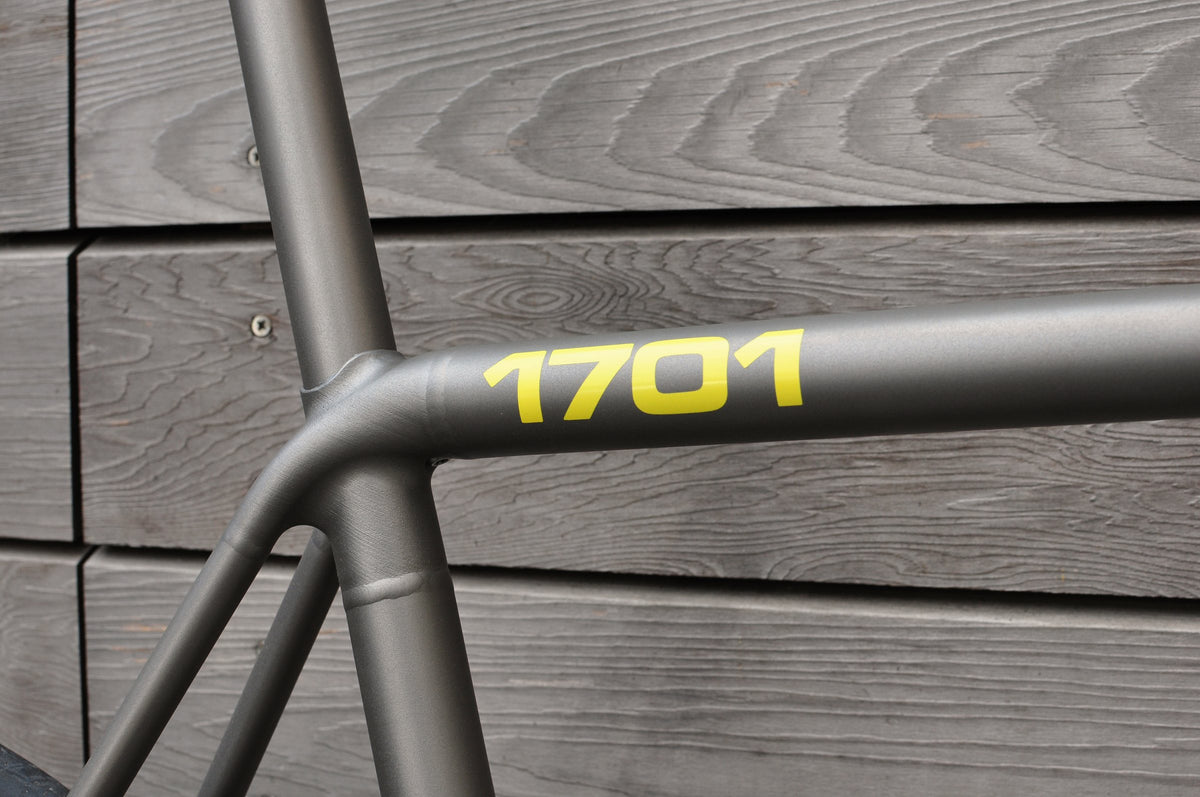 Seat cluster of a titanium frame with the number 1701 in yellow on the top tube, timber cladding behind