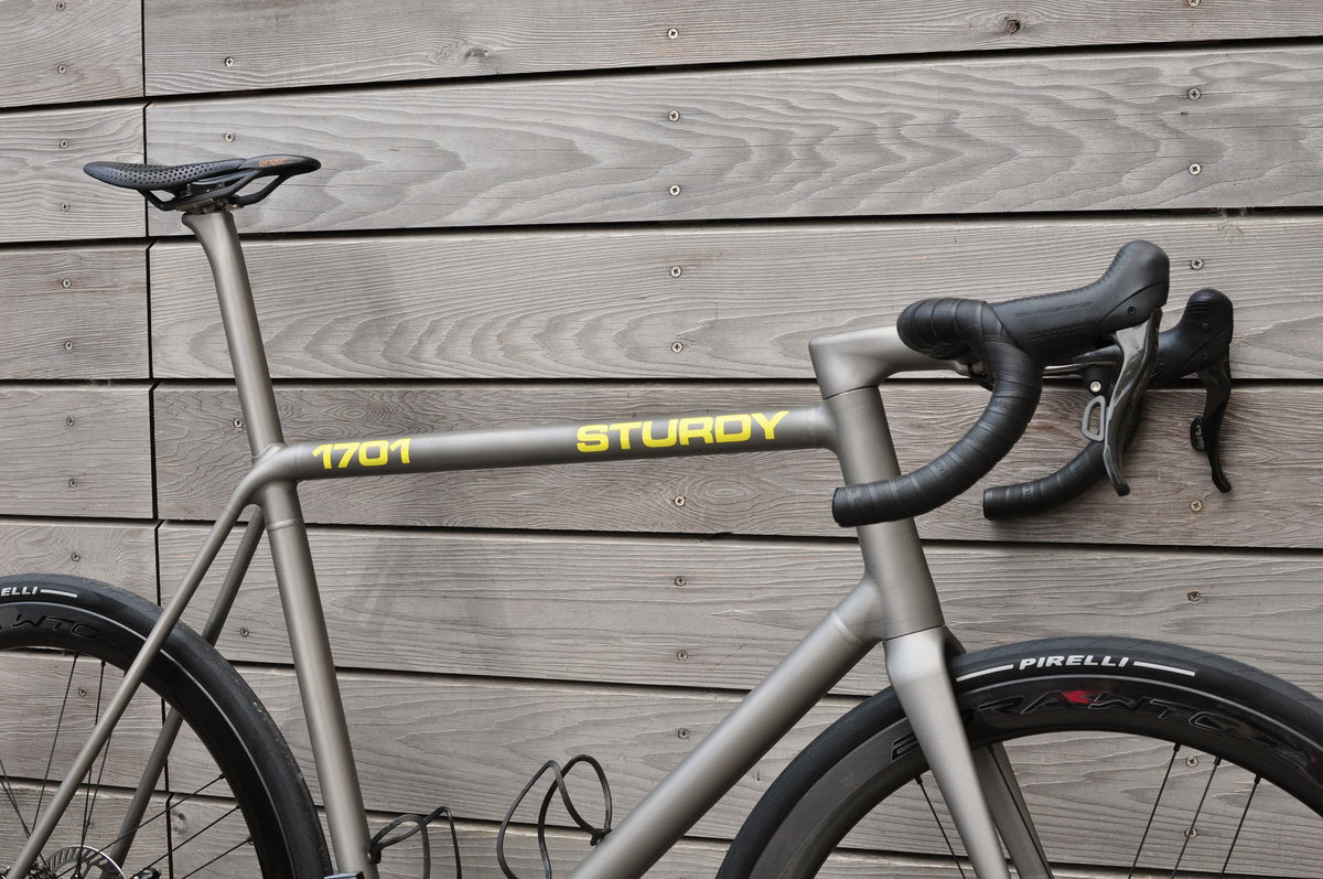 Front half of a titanium road bike with yellow Sturdy and 1701 lettering on the top tube against timber cladding