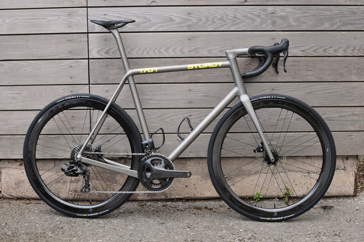 Titanium road bike with yellow Sturdy and 1701 top tube lettering and deep-section wheels against timber cladding