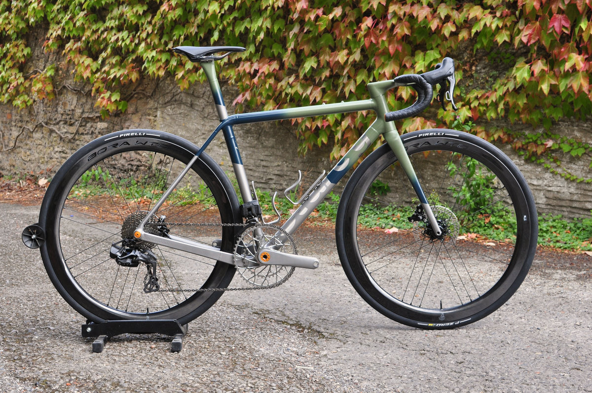 Road bike with a sage green to navy painted fade over titanium, on a stand in front of an autumn ivy wall
