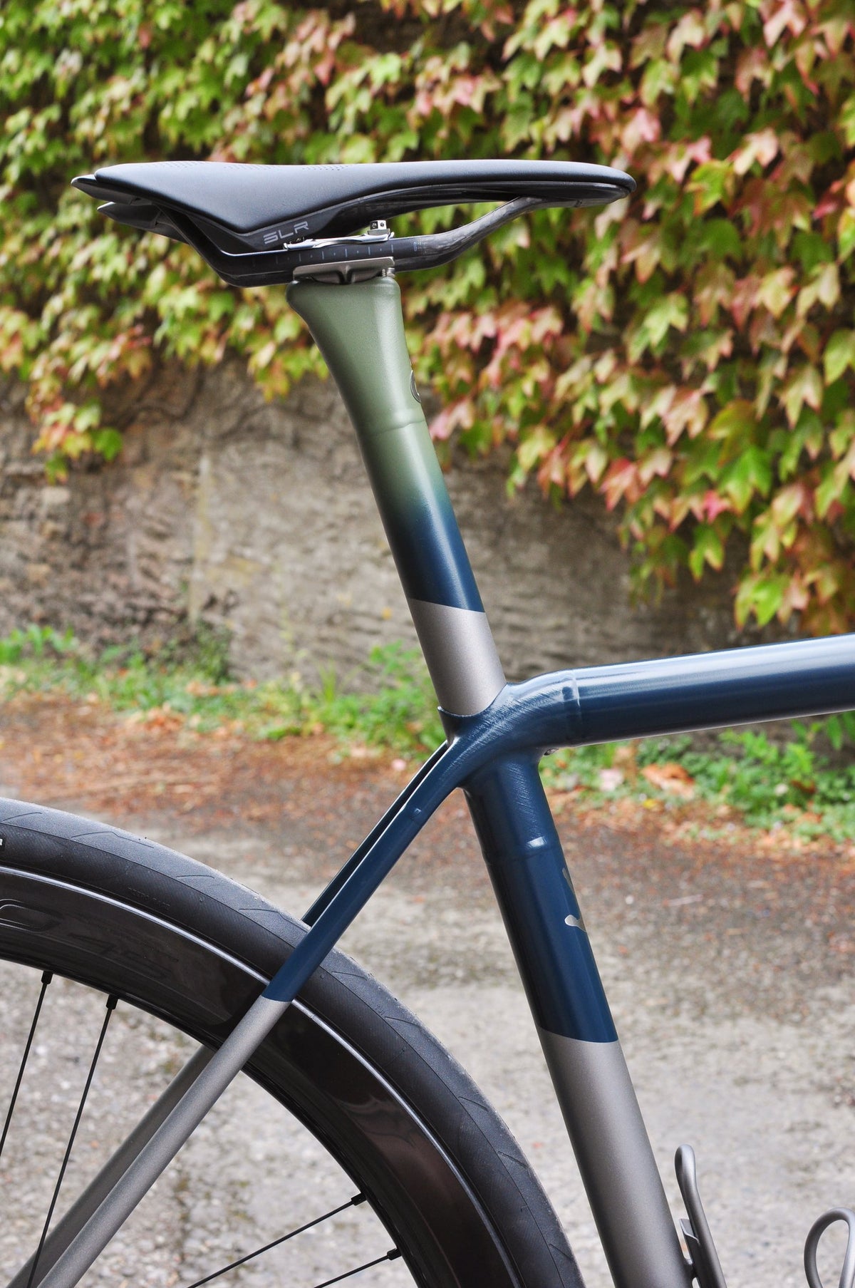 Seat post and seat cluster painted in a sage green to navy fade over bare titanium, ivy-covered wall behind