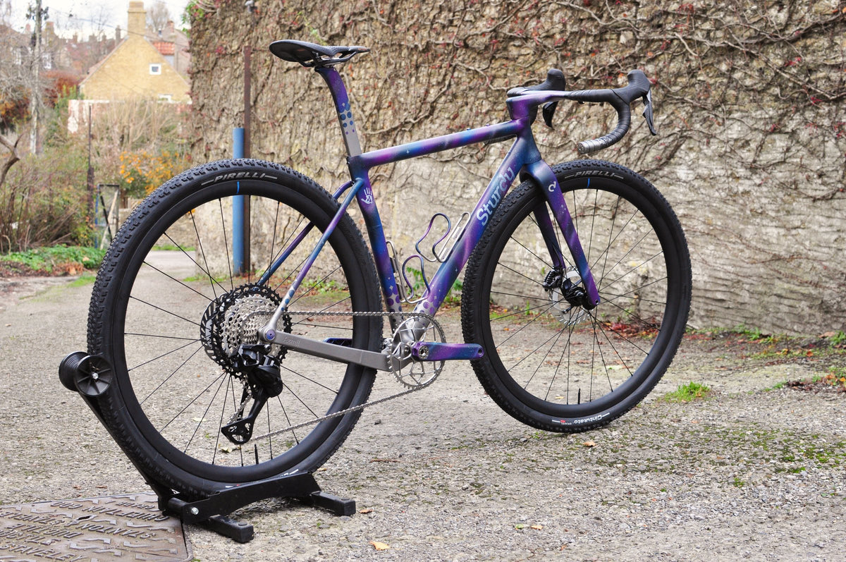 Gravel bike in purple and blue galaxy paint with Sturdy logo and knobbly tyres on a stand by a stone wall