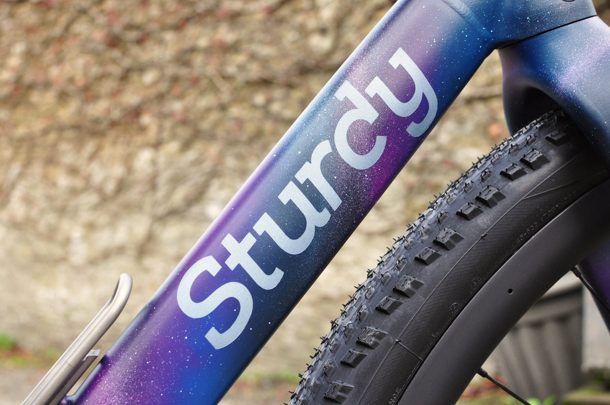 Sturdy logo on a down tube painted in a purple and blue galaxy fade with white speckles, knobbly tyre beside
