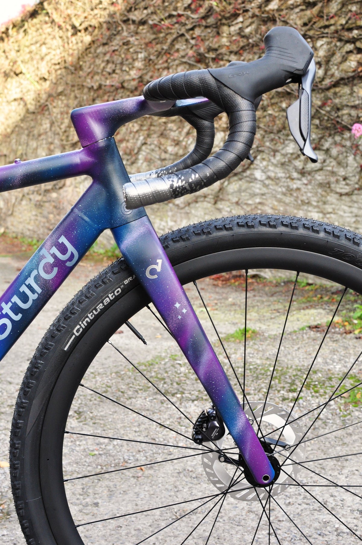 Front end of a gravel bike in purple and blue galaxy paint with Sturdy logo and star motifs, stone wall behind