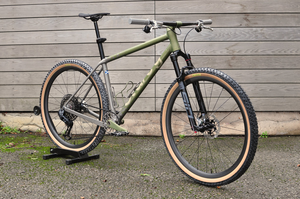 Titanium hardtail mountain bike with olive green paint, tan-wall tyres and Sturdy logo by a timber-clad wall