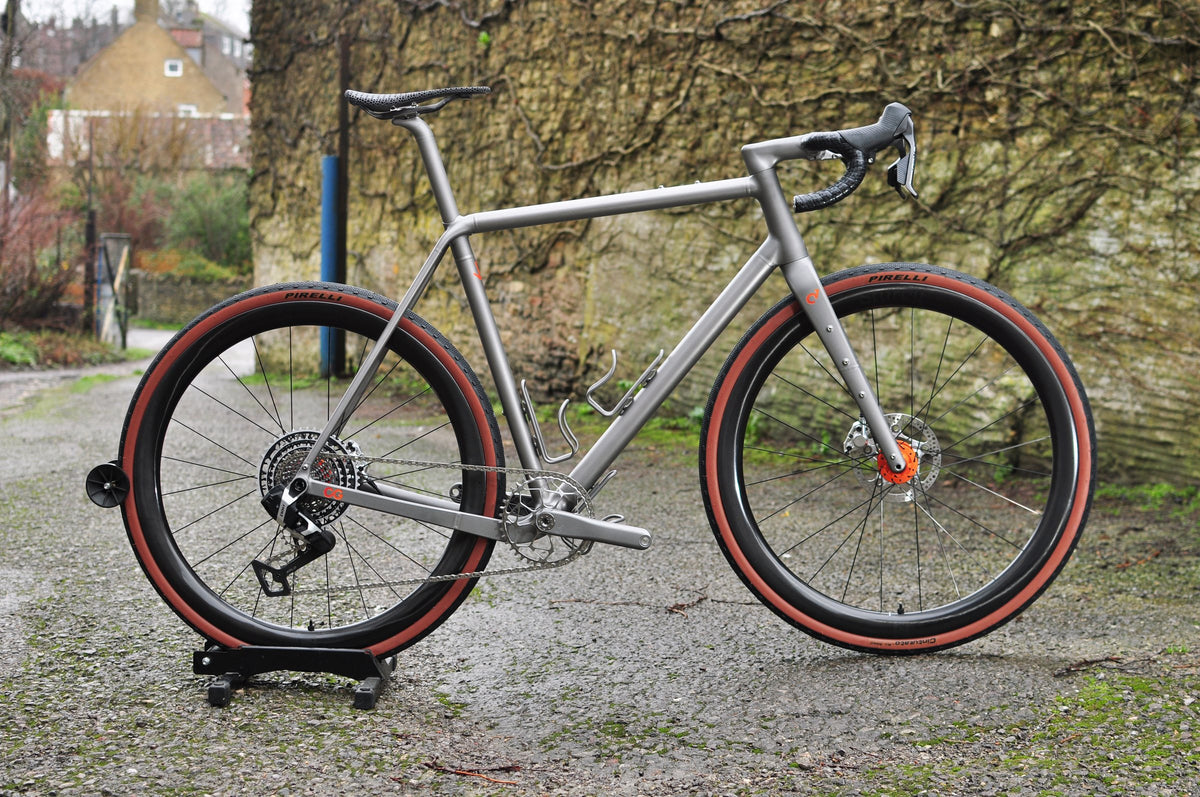 Raw titanium gravel bike with orange details and red-wall tyres on a workstand by an ivy-covered wall