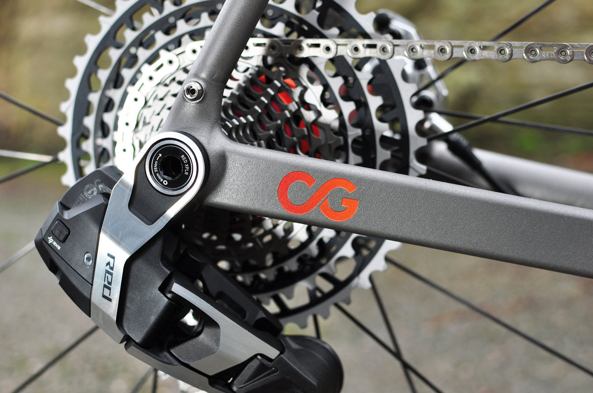 Titanium chainstay with orange initials logo beside a rear derailleur and large cassette