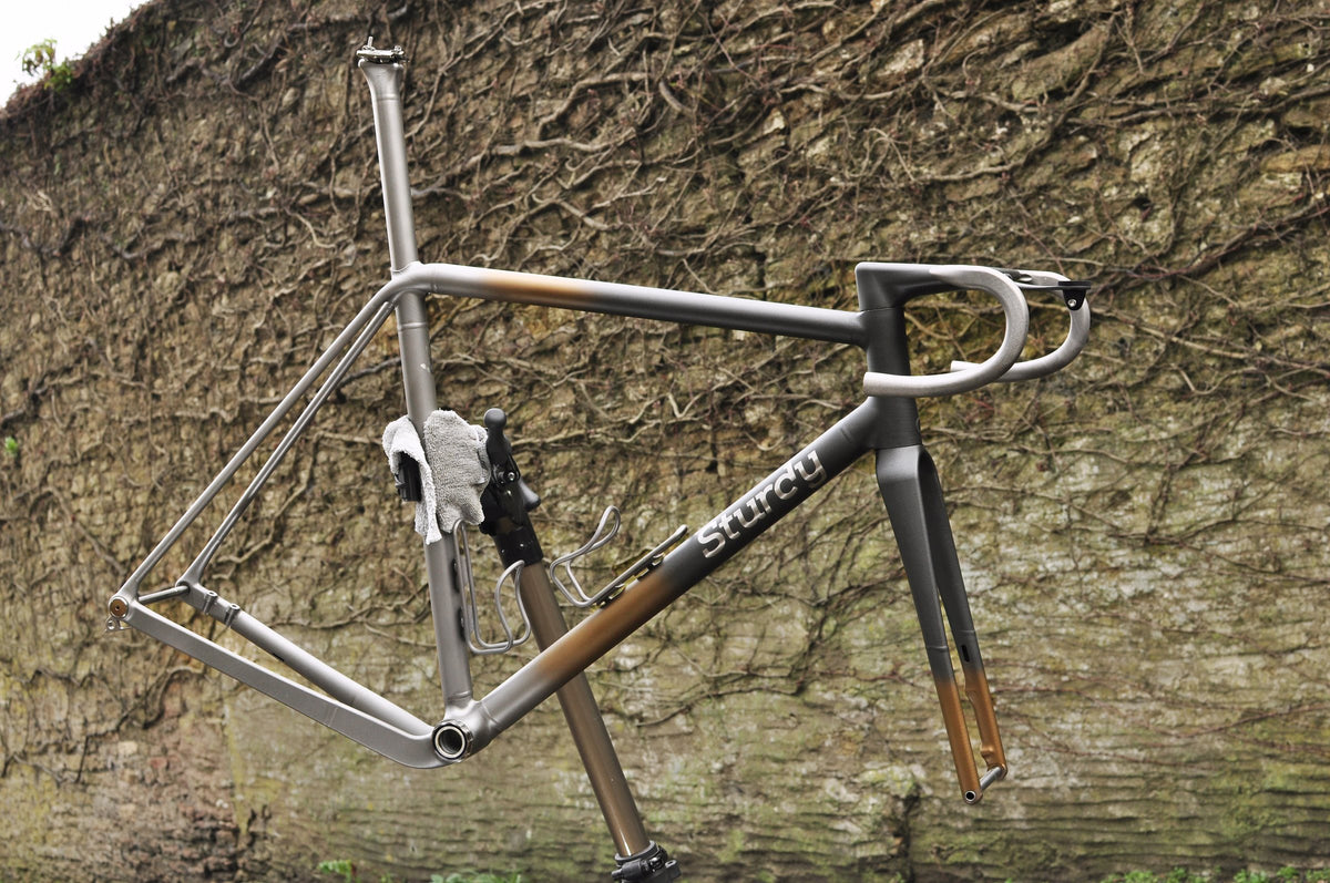 Titanium road frame with dark grey and bronze fade paint and Sturdy logo on a stand by a stone wall