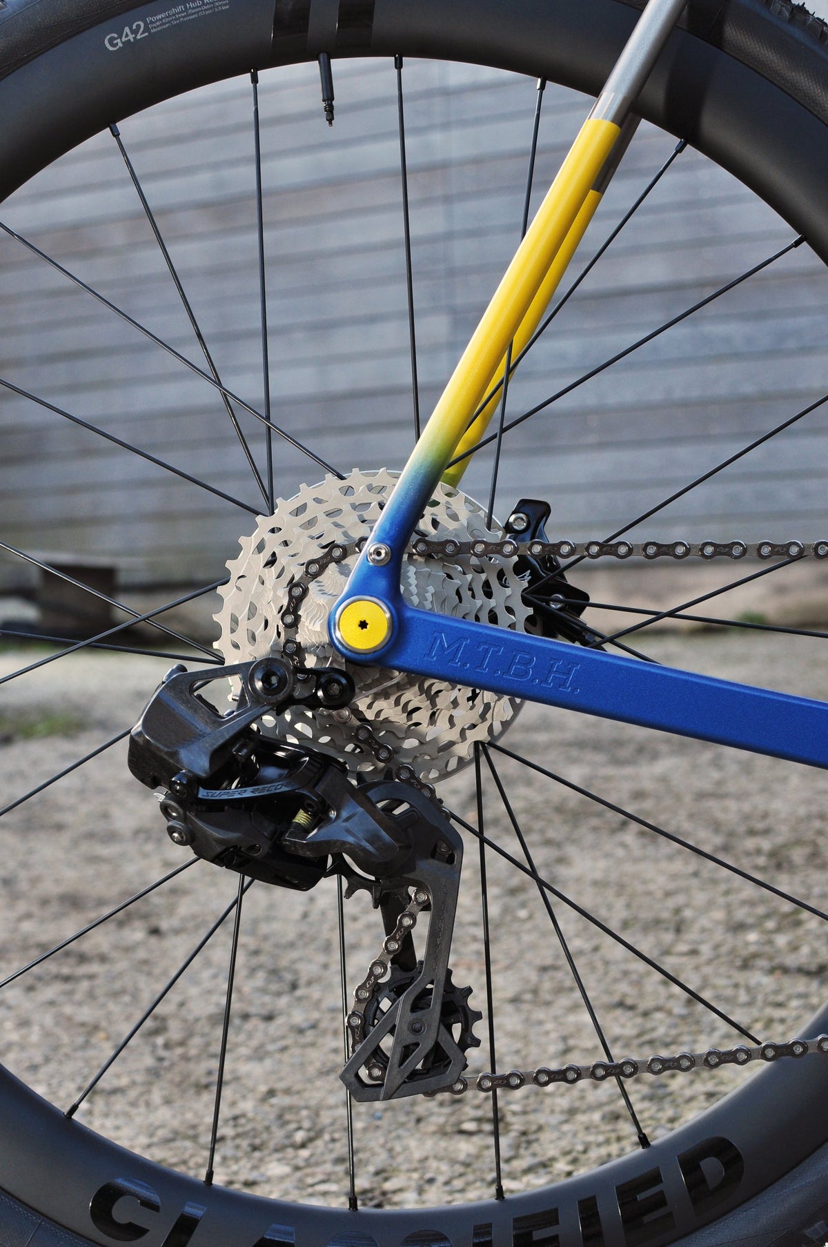 Yellow-to-blue painted rear stays with custom lettering beside a cassette and rear derailleur