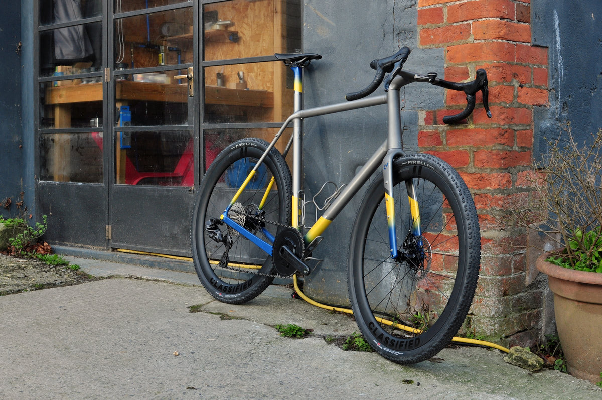 Titanium gravel bike with yellow and blue painted details leaning outside a workshop door