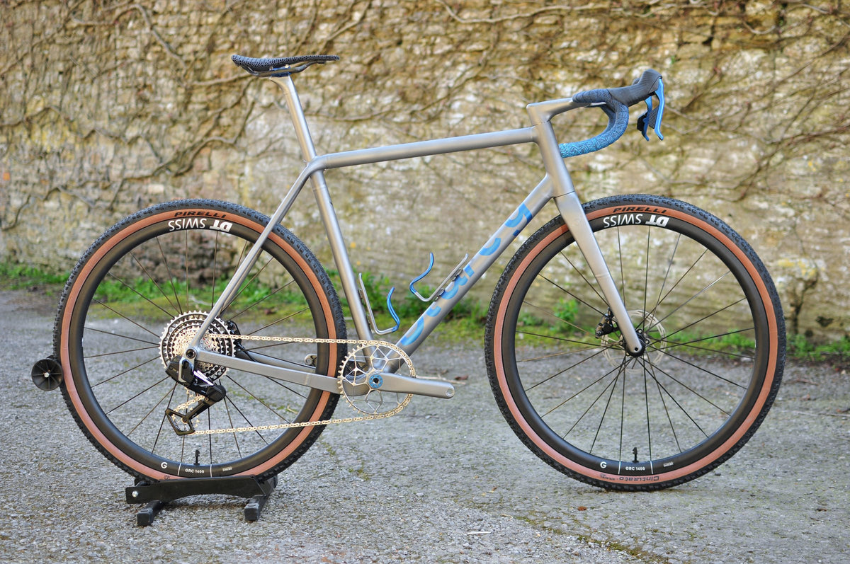 Titanium gravel bike with blue Sturdy logo, blue bar tape and tan-wall tyres by a stone wall