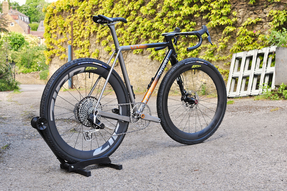 Titanium gravel bike with black-to-orange fade front triangle and deep-section wheels on a stand in a lane