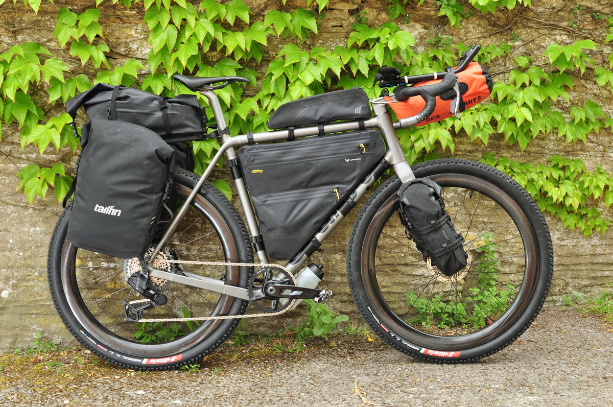 Titanium gravel bike loaded with black panniers, frame bag and orange bar roll against an ivy-covered wall