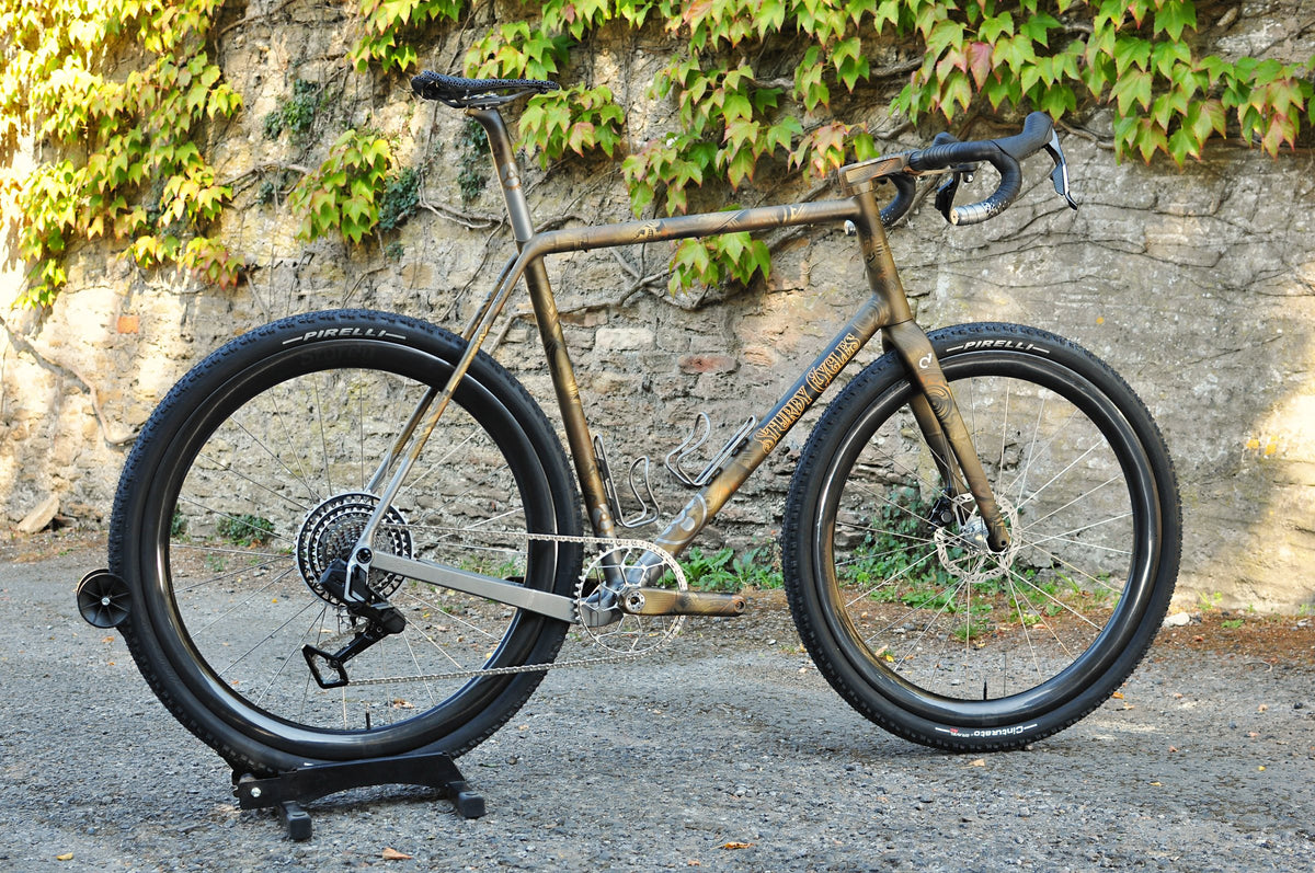 Gravel bike in bronze and gold tribal-pattern paint with ornate Sturdy Cycles lettering by an ivy-covered wall