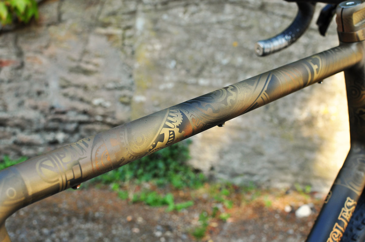 Bronze top tube with a swirling tribal-style masked pattern against a blurred stone wall