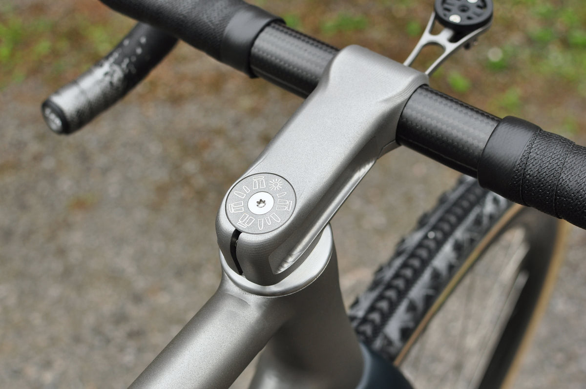 Titanium stem with engraved top cap and carbon handlebar, viewed from above over a gravel tyre