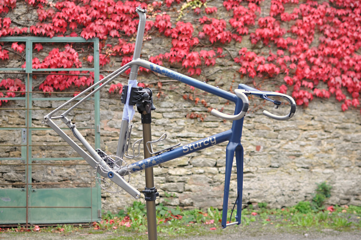 Navy blue and titanium frameset with Sturdy logo and hexagon fade in a workstand by red ivy