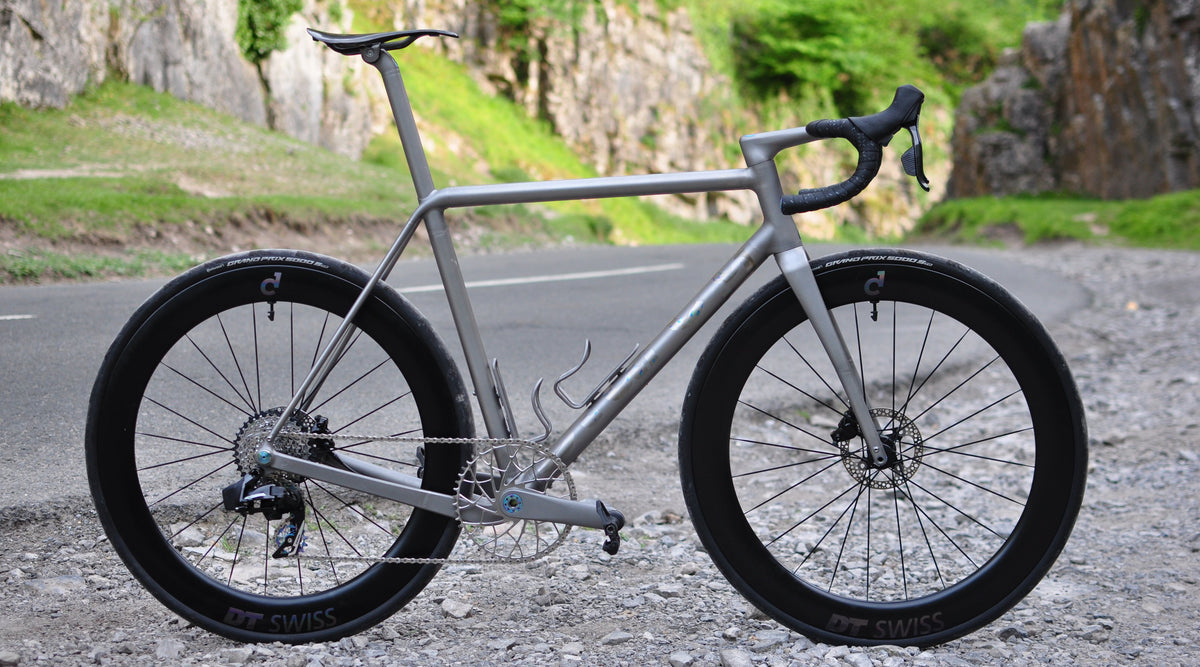 Raw titanium road bike with skeletal chainring and deep carbon wheels on a gravel verge in a limestone gorge